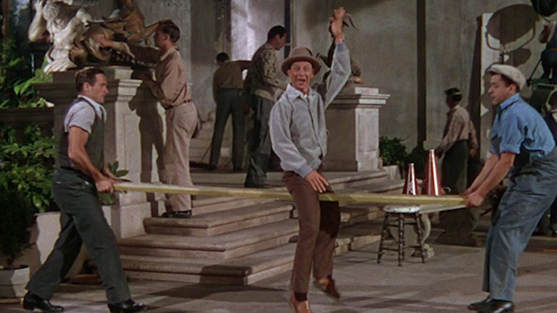 Donald O'Connor as Cosmo sitting on wood plank being carried by two men with hand up in Singing in the Rain