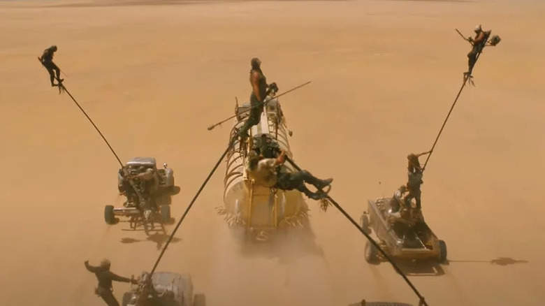 Four men (faces not visible) hanging atop long poles attached to souped-up cars chasing a large tanker in Mad Max: Fury Road
