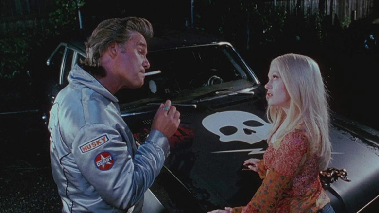 Kurt Russell as Stuntman Mike talking to Rose McGowan as Pam in front of his black car with a skull and crossed lightning bolts on the hood in Death Proof