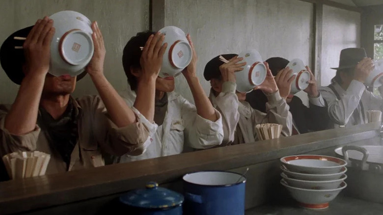 A row of men sitting at counter drinking from ramen bowls covering their faces in Tampopo