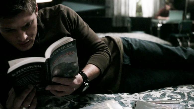Dean lies on bed reading Supernatural book