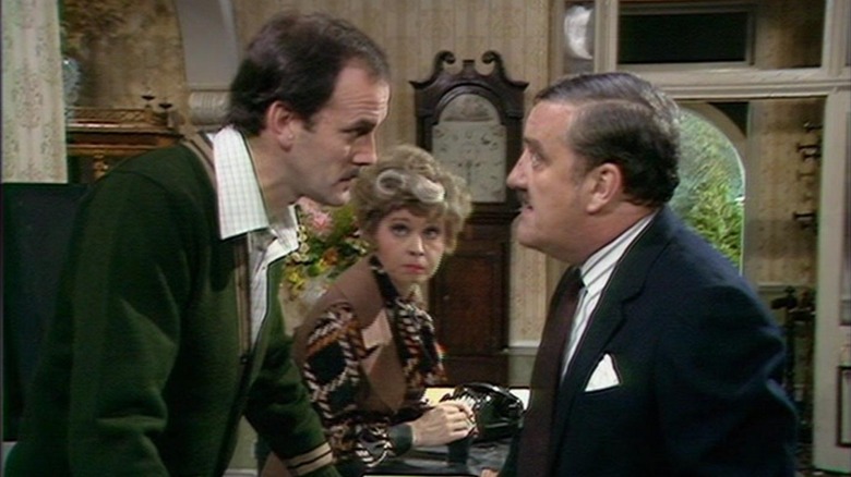 Basil Fawlty (John Cleese), Sybil Fawlty (Prunella Scales), and Mr. Hutchinson (Bernard Cribbins) bicker over hotel matters in Fawlty Towers