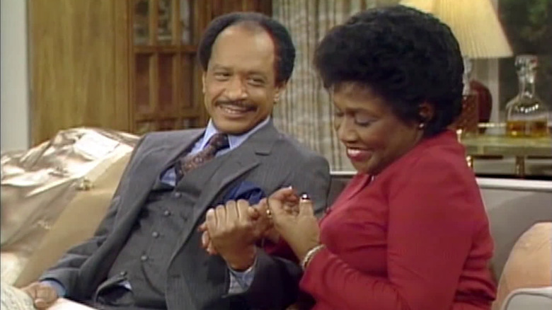George Jefferson (Sherman Hemsley) and Weezey Jefferson (Isabel Sanford) hold hands and laugh in The Jeffersons