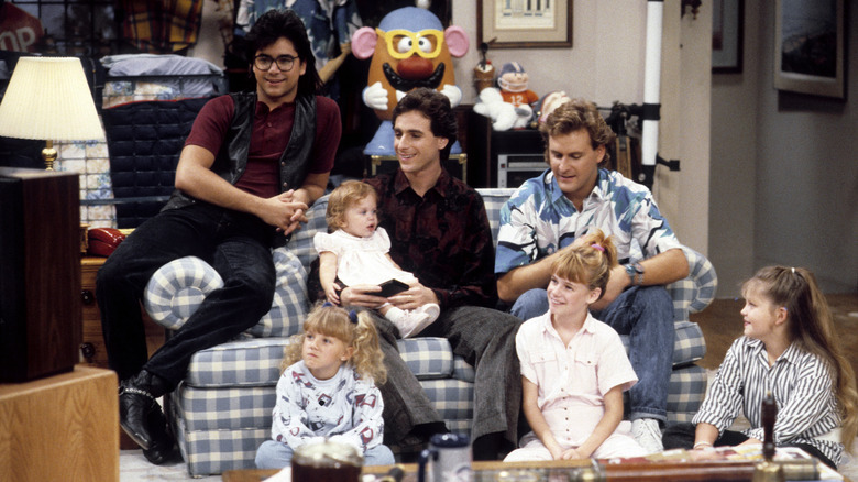 John Stamos, Bob Saget, Mary-Kate/Ashley Olsen, Dave Coulier, Jodie Sweetin, Andrea Barber, and Candice Cameron Bure as the Tanner family (and Kimmy) watching TV in the living room in Full House