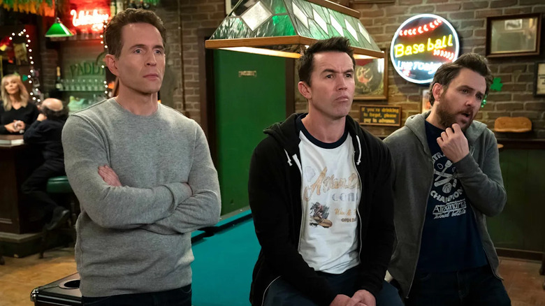 Glenn Howerton as Dennis Reynolds, Rob McElhenny as Mac McDonald, and Charlie Day as Charlie Kelly stand in Paddy's Pub in It's Always Sunny in Philadelphia.