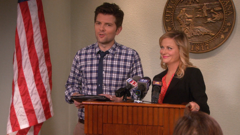 Adam Scott as Ben Wyatt stands at a podium next to Amy Poehler as Leslie Knope in Parks and Recreation.