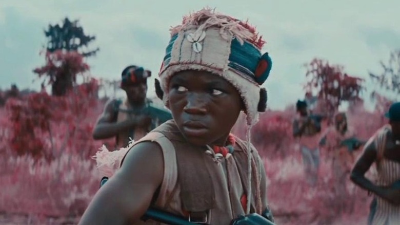 Abraham Attah as Agu standing in a pink grass field and holding a gun in Beasts of No Nation