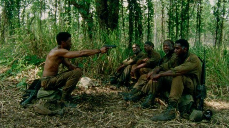 Chadwick Boseman as Stormin' Norman, Delroy Lindo as Paul, Clarke Peters as Otis, Isiah Whitlock Jr. as Melvin, and Norm Lewis as Eddie sitting in the woods while Norman points a gun at the rest in Da 5 Bloods