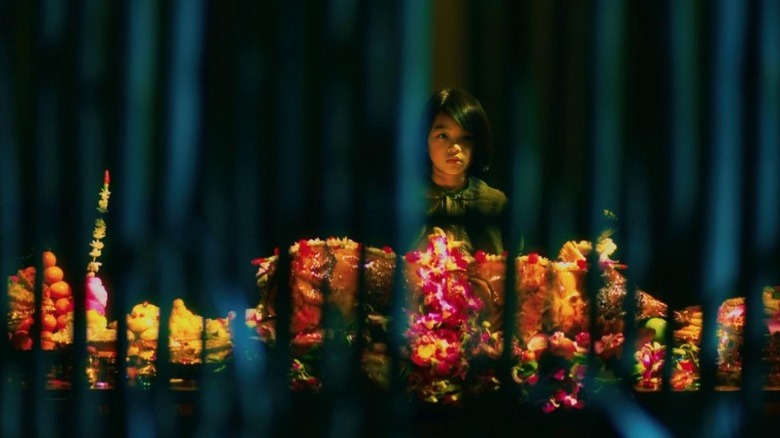 Sareum Srey Moch as Loung Ung standing in the dark behind a table in First They Killed My Father