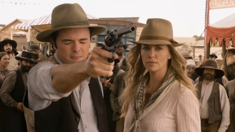 Seth MacFarlane as Albert Stark pointing a gun with his eyes closed while Charlize Theron as Anna Barnes-Leatherwood gazes sternly at his target in A Million Ways to Die in the West