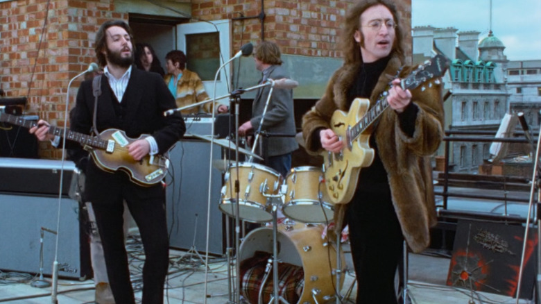 Paul McCartney stands next to John Lennon as they play music on a rooftop in The Beatles: Get Back