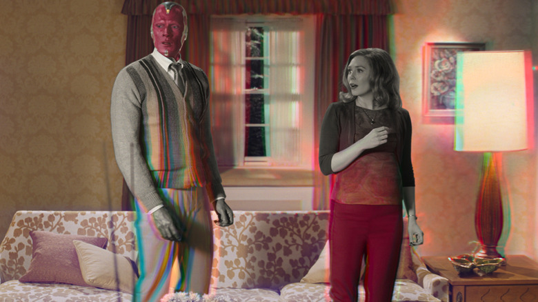 Paul Bettany as Vision stands in a living room with Elizabeth Olsen as Wanda Maximoff as the screen changes colors in WandaVision