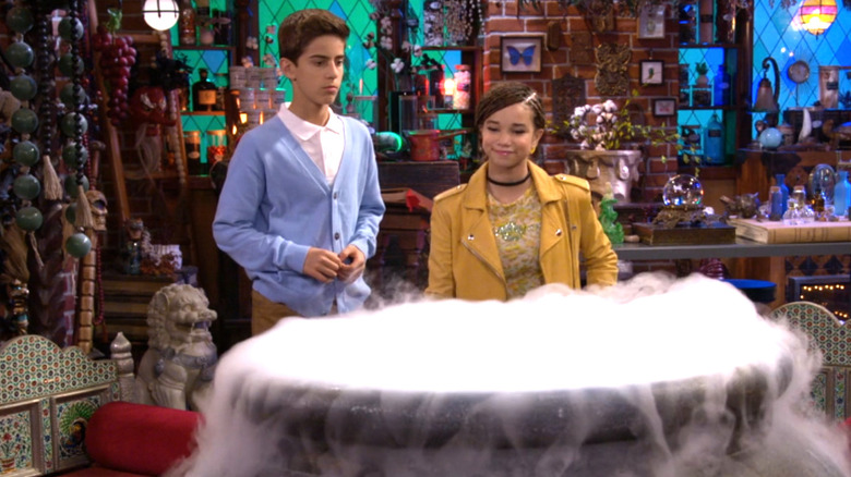 Alkaio Thiele as Roman Russo stands behind a cauldron with Janice LeAnn Brown as Billie in Wizards Beyond Waverly Place