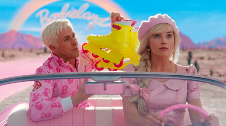 Ryan Gosling as Ken in a pink jacket holding up neon rollerblades in the backseat of a car driven by Margot Robbie as Barbie away from Barbieland in Barbie