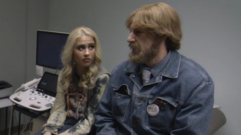Maria Bakalova as Tutar in a blonde wig sitting next to Sacha Baron Cohen as Borat in disguise as a fat American man in a doctor's office in Borat Subsequent Moviefilm