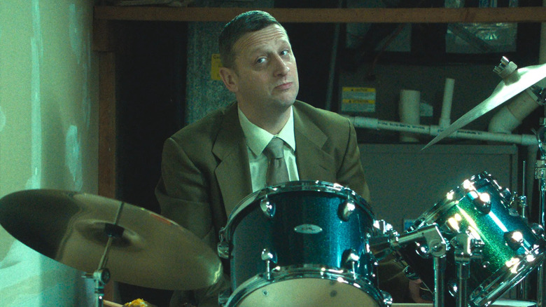 Tim Robinson as Craig wearing a suit and sitting behind a drum kit in his garage in Friendship