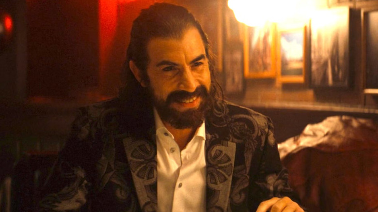 Sacha Baron Cohen as Mephisto wearing a suit in a restaurant and smiling in Ironheart