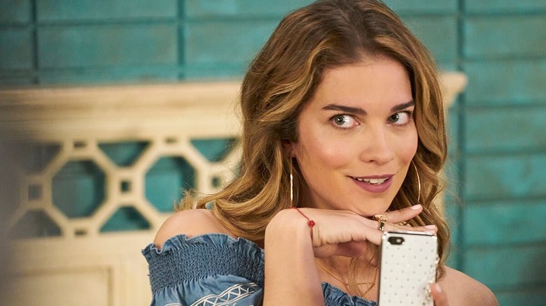 Alexis holds her phone in one hand, with the other under her chin, smiling mischievously