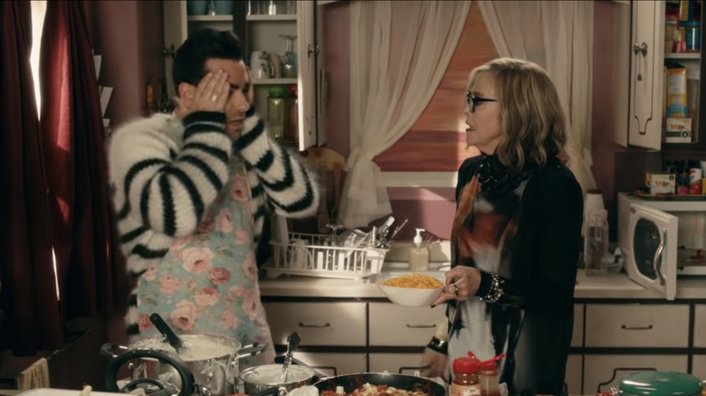 David covers his face with his hands as Moira looks at him, holding a bowl of cheese