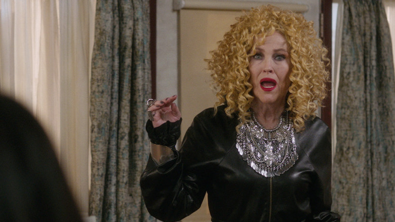 Mora wears a dramatic gown and big curly wig, looking on with an open mouth