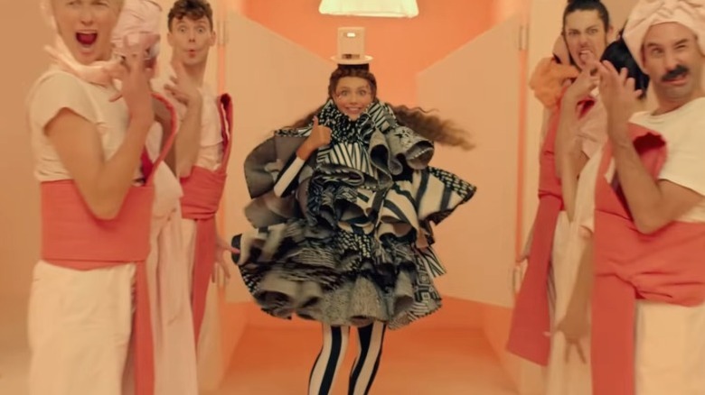 Maddie Ziegler as Music wearing a multi-layered black-and-white garment and giving a thumbs up while surrounded by dancers in Music