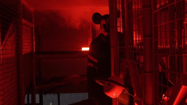 Simon Phillips as Tim Collins wearing a Mickey Mouse mask and standing behind a row of metal cages in The Mouse Trap