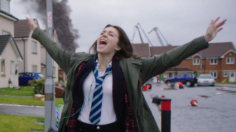 Anna walks down street with headphones in, oblivious to zombies behind her