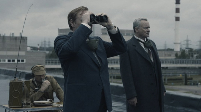 Standing on a rooftop, Legasov looks through binoculars while Shcherbina stands next to him and looks out over a city in Chernobyl
