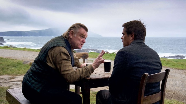 Colm and Padraic sit at a table outside, with Colm looking fiercely and holding up his right hand, which is missing one finger.