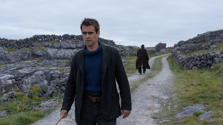 Padraic walks down a dirt road on a cloudy day, with a pensive look on his face, as a man walks in the opposite direction behind him.