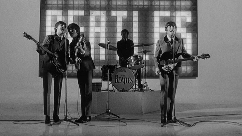 three men playing guitars in suits and a fourth man playing drums in front of a "beatles" backdrop