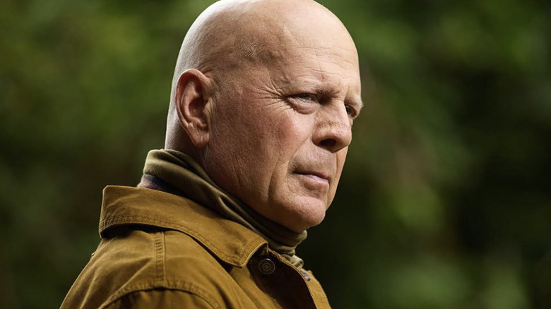 Bruce Willis as Robert stands alone outside in a brown jacket against a green forest backdrop in Fortress