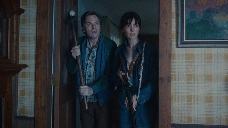 Annie Hathaway as Denise Platt and Ewan McGregor as Greg Platt in The End of Oak Street, standing in a doorway as Greg wields a sledgehammer and Denise wields a rifle.