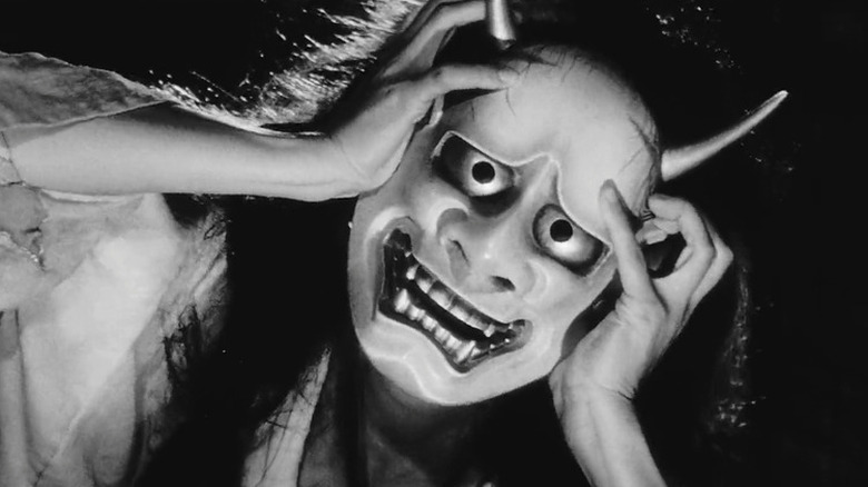 A woman wears a samurai mask with a haunting grin.