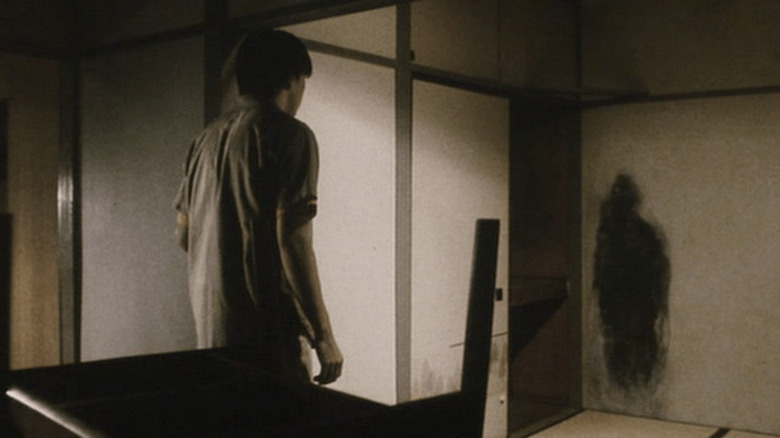 A man stands with his back to the camera as he looks at a human-shaped stain on his wall.