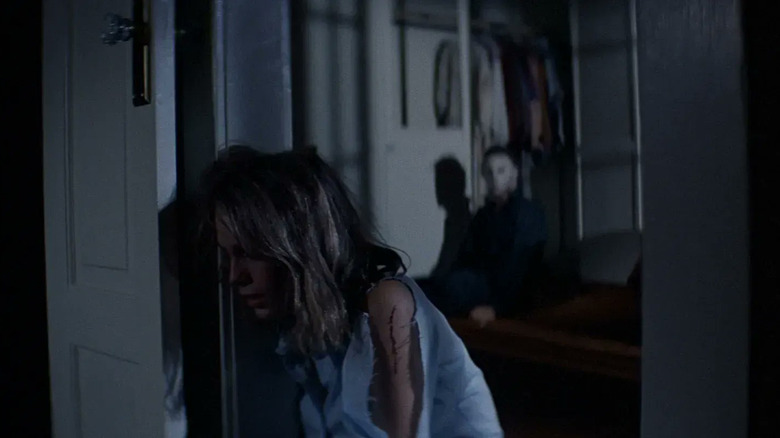 Laurie (Jamie Lee Curtis) leans against a door frame as Michael Myers (Nick Castle) sits up in the background in Halloween (1978)