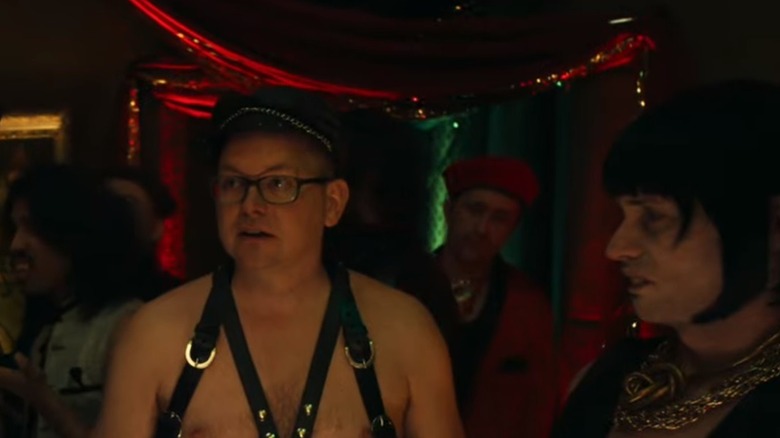 Colin stands at a party in a leather harness