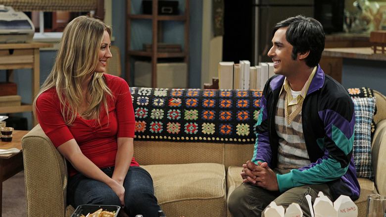 Penny and Raj sitting on her couch smiling and talking on The Big Bang Theory