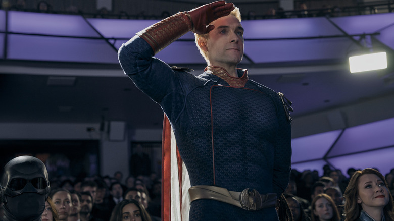 Antony Starr as Homelander standing up amid a large crowd of seated onlookers and saluting in The Boys
