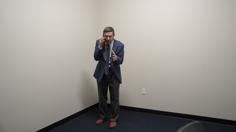 Ron stands in a corner and talks on the phone in The Chair Company