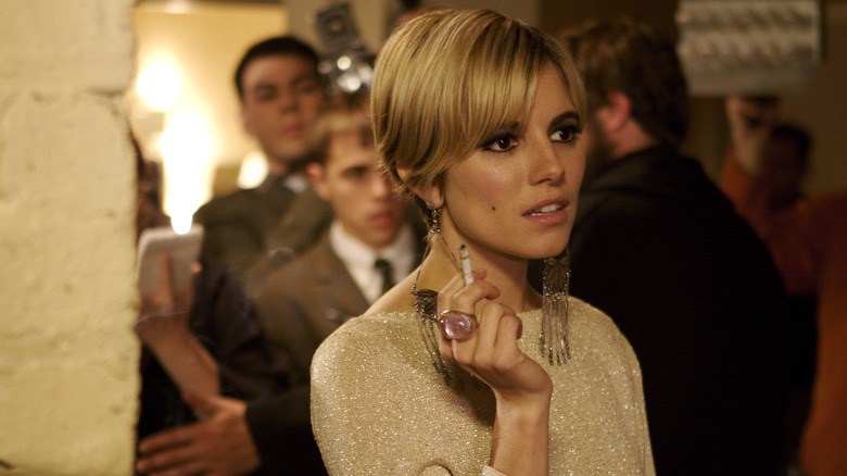 blonde woman with long earrings holding a cigarette with several men behind her