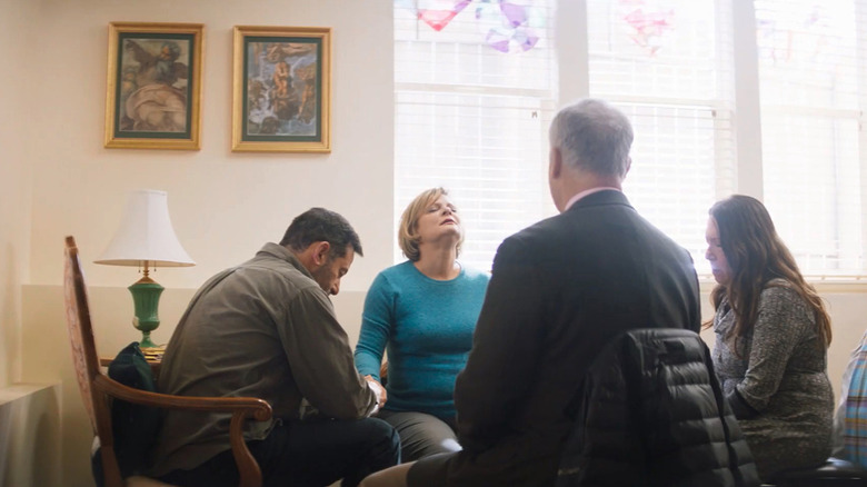 two men and two women sitting in a circle near a window with one of the women with her head back and eyes closed
