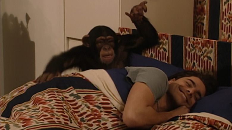 John Stamos dans le rôle de l'oncle Jesse dans Full House, allongé dans son lit, souriant avec un chimpanzé derrière lui, le frappant à l'arrière