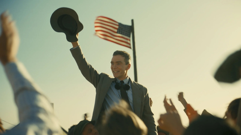 J. Robert Oppenheimer raises his hat in celebration for a cheering audience with the American flag waving behind him in 