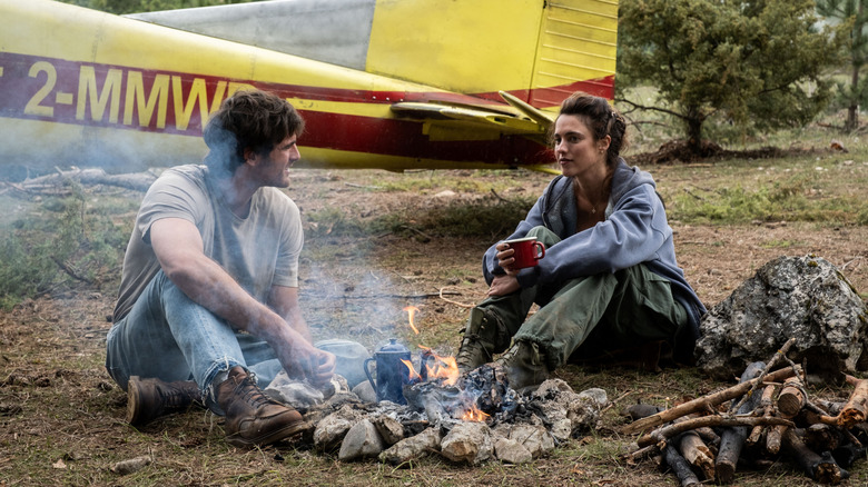 Jacob Elordi as Hig and Margaret Qualley as Cima warming themselves by a campfire next to a small plane in The Dog Stars