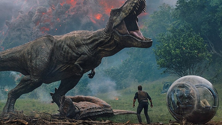 A T-Rex roars as it stands on top of a Carnotaurus behind a gyrosphere in Jurassic World: Fallen Kingdom