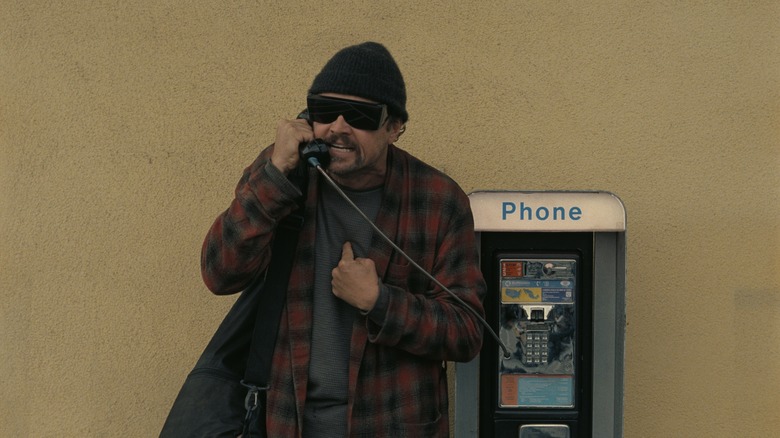 Bob talking on a payphone in a hat, sunglasses, and red bathrobe in One Battle After Another