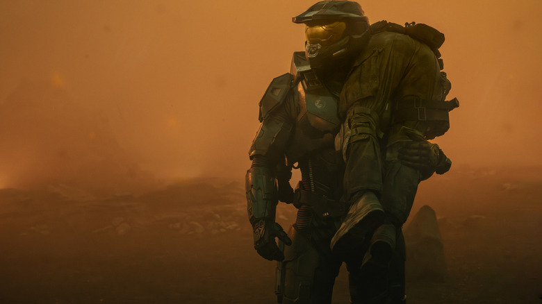 Pablo Schreiber as Master Chief standing on an alien world covered in fog and carrying a fallen soldier over his shoulder in Halo
