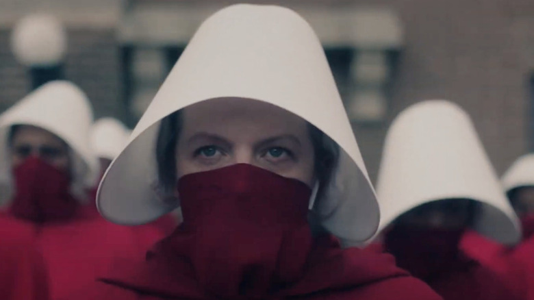 June (Elisabeth Moss) wears her red Handmaid's cloak, white bonnet, and a red face covering pulled up to her nose, standing among other Handmaids and staring ahead with an intense gaze in The Handmaid's Tale