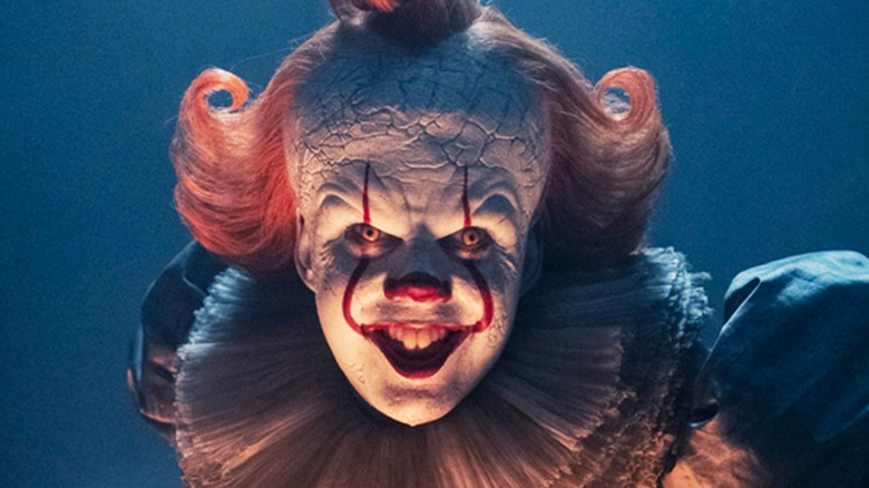 Bill Skarsgård's Pennywise grins as he crouches in a dark room in It: Welcome to Derry